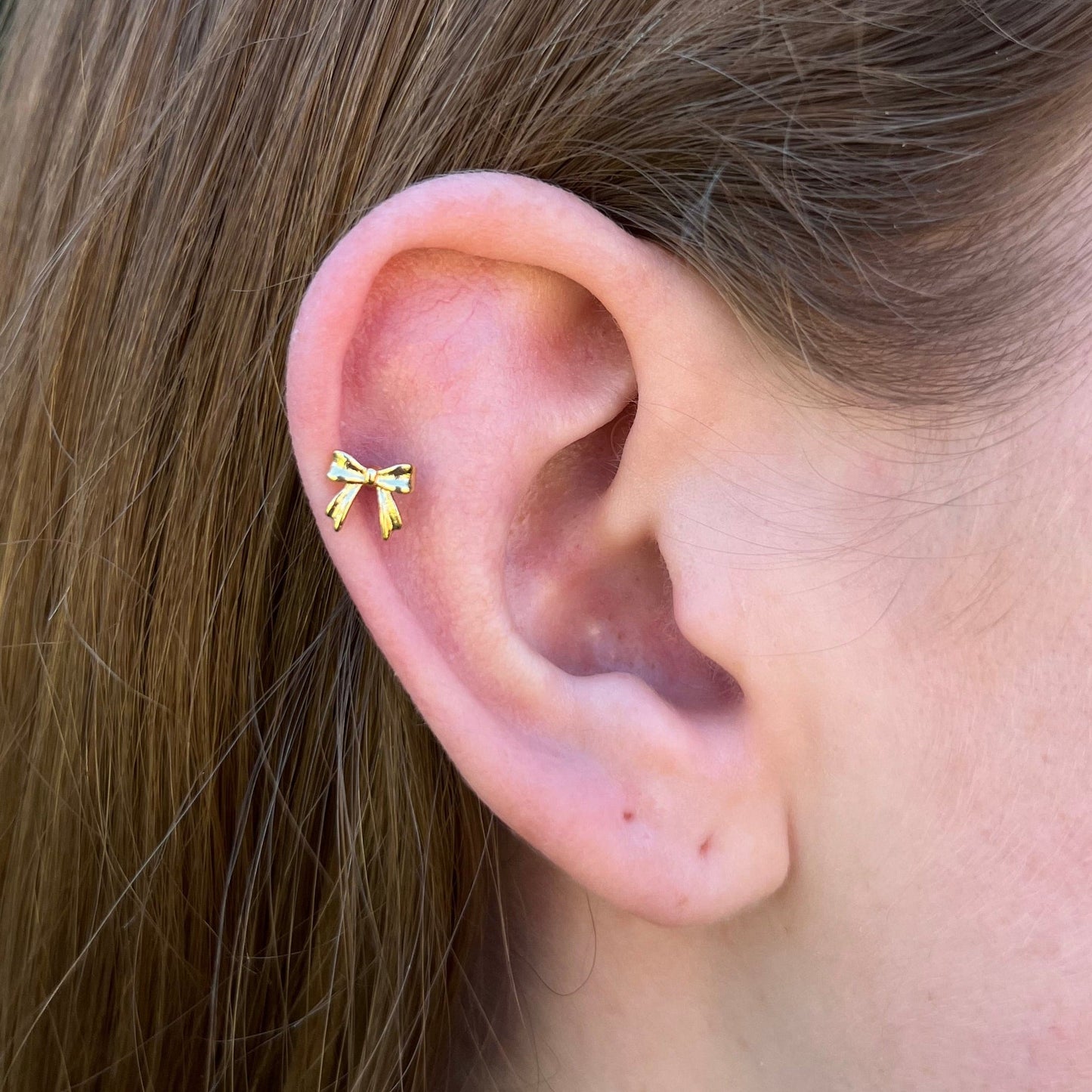 Close-up of an ear wearing a gold bow flatback earring with a blurred green background.